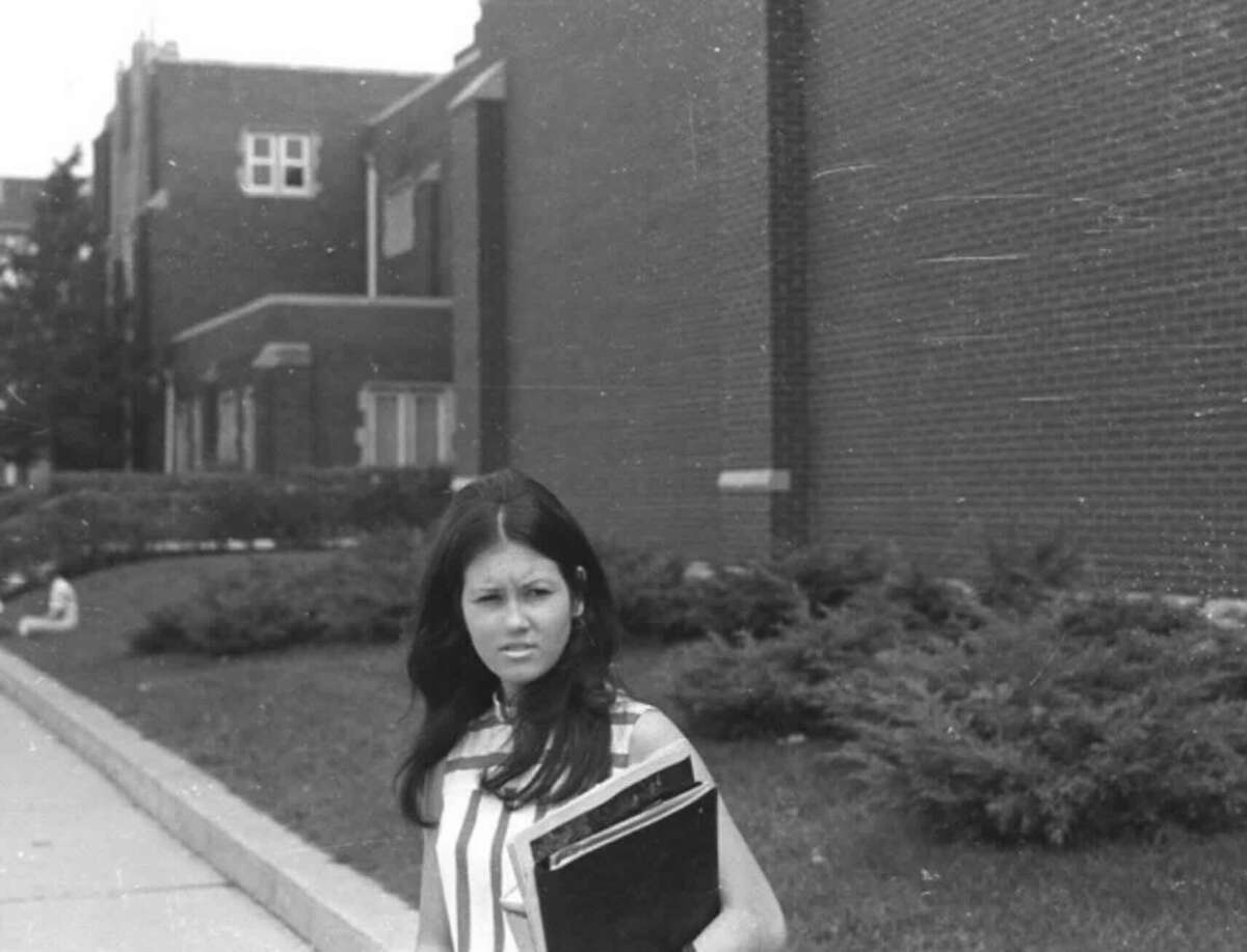 A young woman with long dark hair holding books stands on a sidewalk next to a brick building, looking ahead with a serious expression. Bushes and another building are visible in the background. The image is black and white.