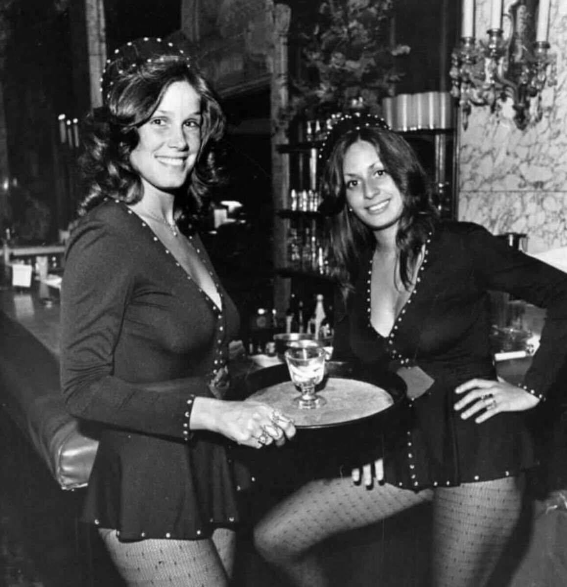 Two women in matching black uniforms with deep necklines and fishnet stockings stand behind a bar. One holds a tray with a cocktail; both are smiling. The bar has ornate decor and bottles lined up in the background.
