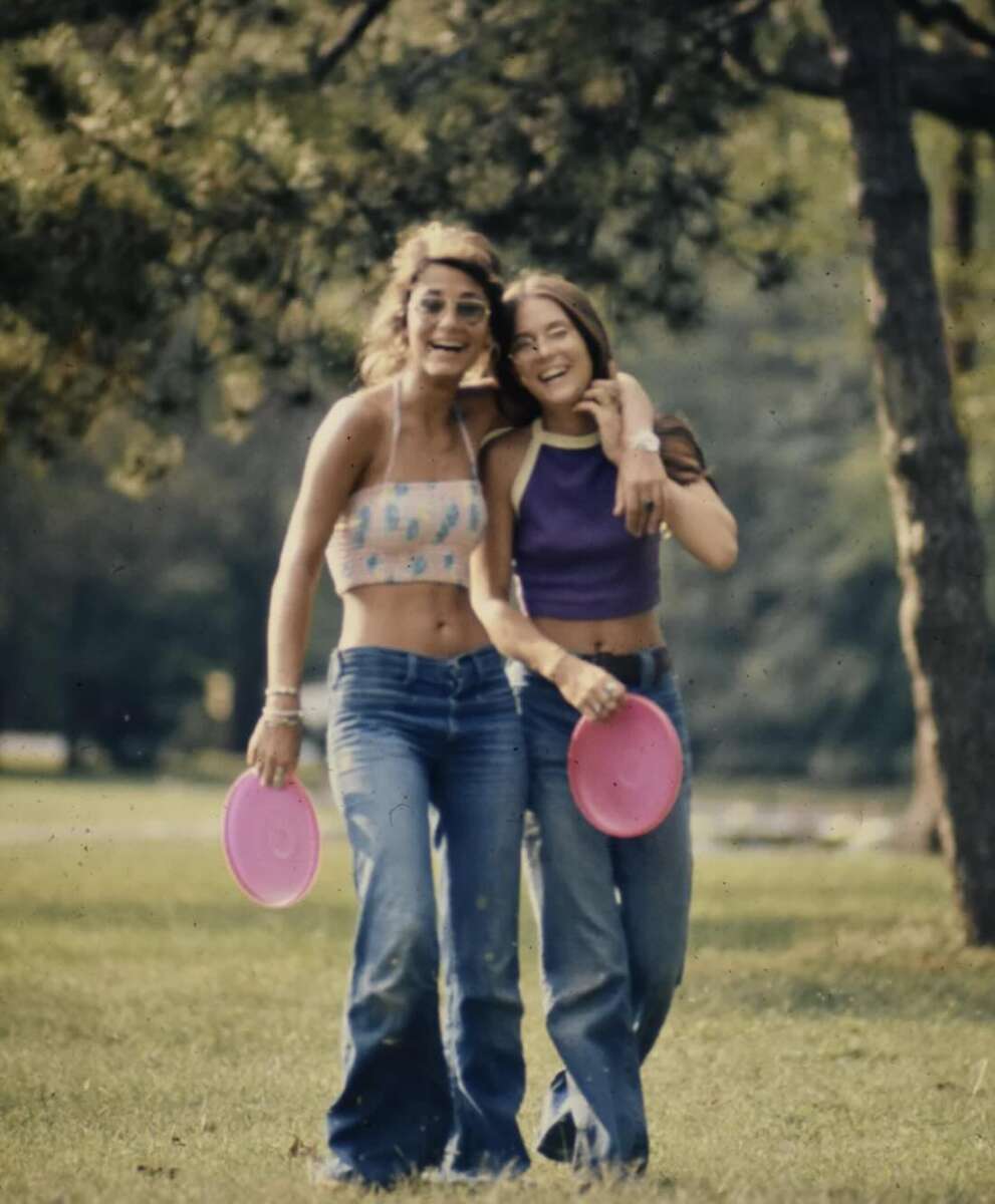 Two young women in casual clothes walk arm-in-arm on grass, smiling and holding pink frisbees, with trees and greenery in the background.