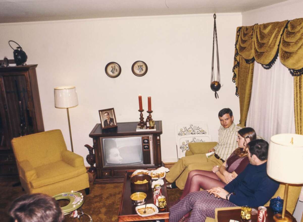 Three people sit talking in a retro living room with gold curtains, a yellow chair, and an old TV. Snacks and drinks are on the coffee table; a portrait and candles sit atop the TV. The decor is vintage, likely from the 1970s.