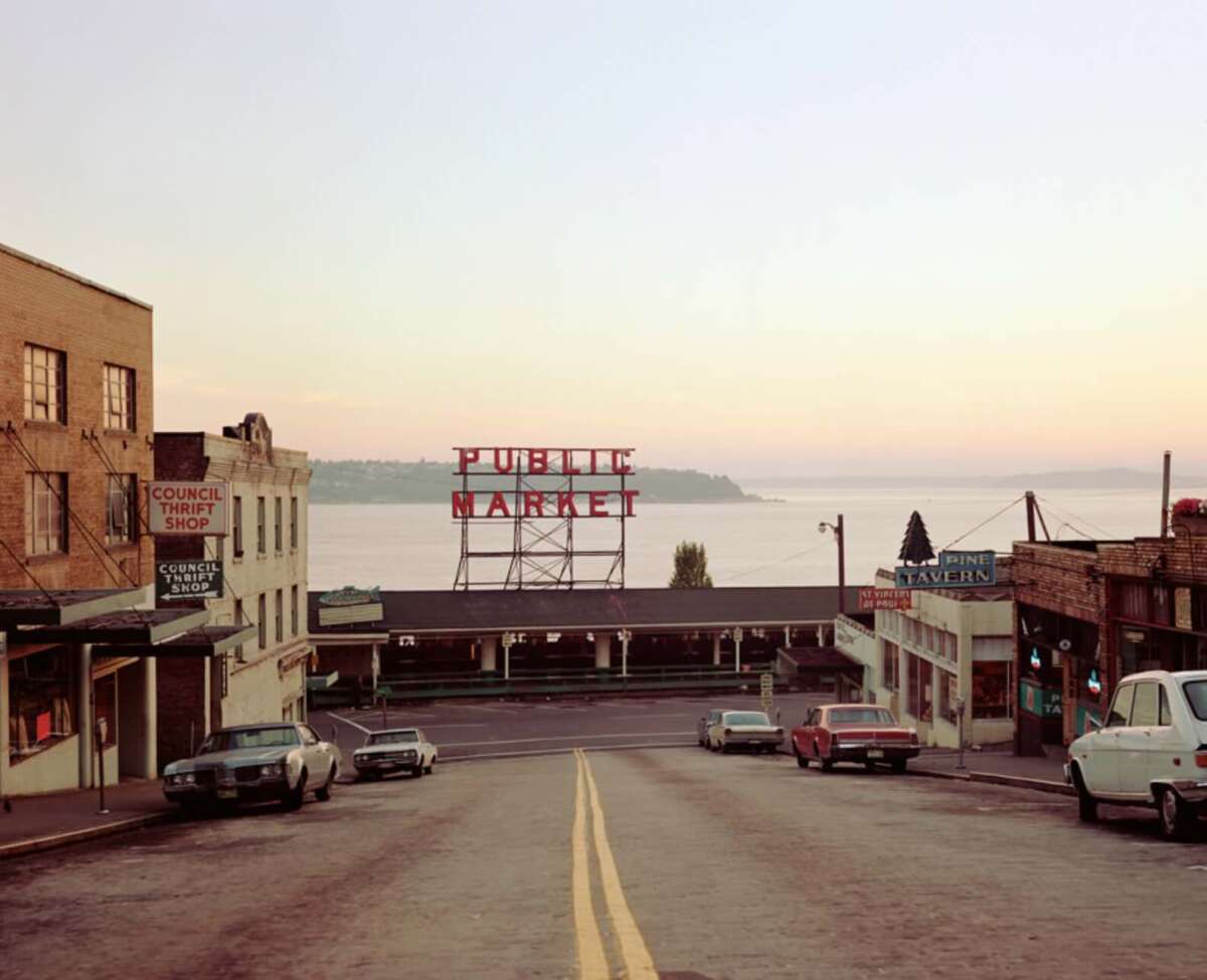 A quiet street slopes down toward Seattle’s iconic Public Market sign at sunset, with vintage cars parked along the sides and storefronts lining the roadway, overlooking calm water in the background.