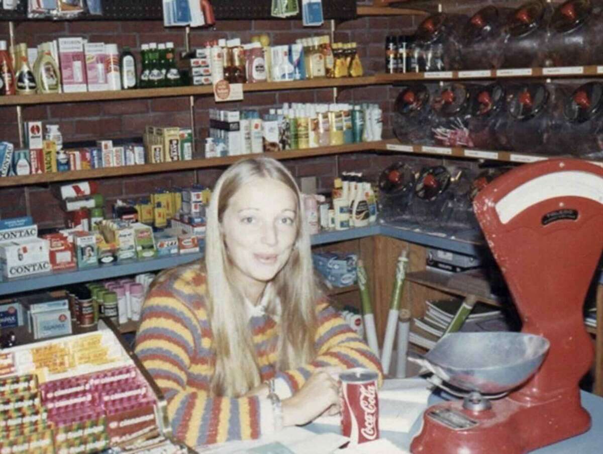 A young woman with long blonde hair, wearing a striped sweater, sits behind a counter in a vintage store, holding a can of Coca-Cola. Behind her are shelves stocked with various products and a red scale on the counter.