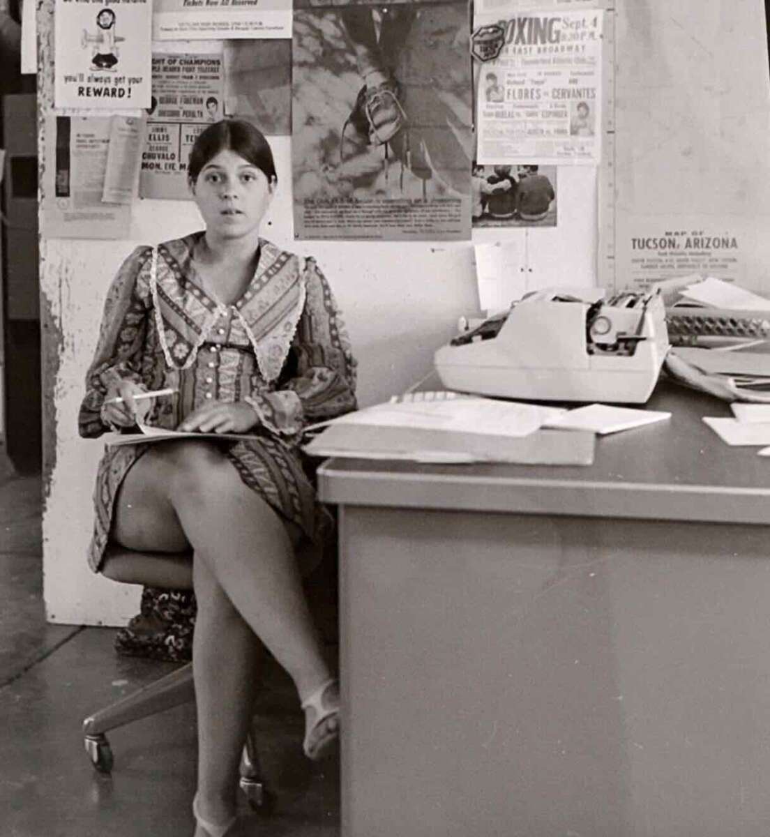 A woman in a patterned dress sits on a chair beside a desk with papers and a typewriter, holding a cup. Behind her, a wall is covered with posters, clippings, and a boxing event flyer.