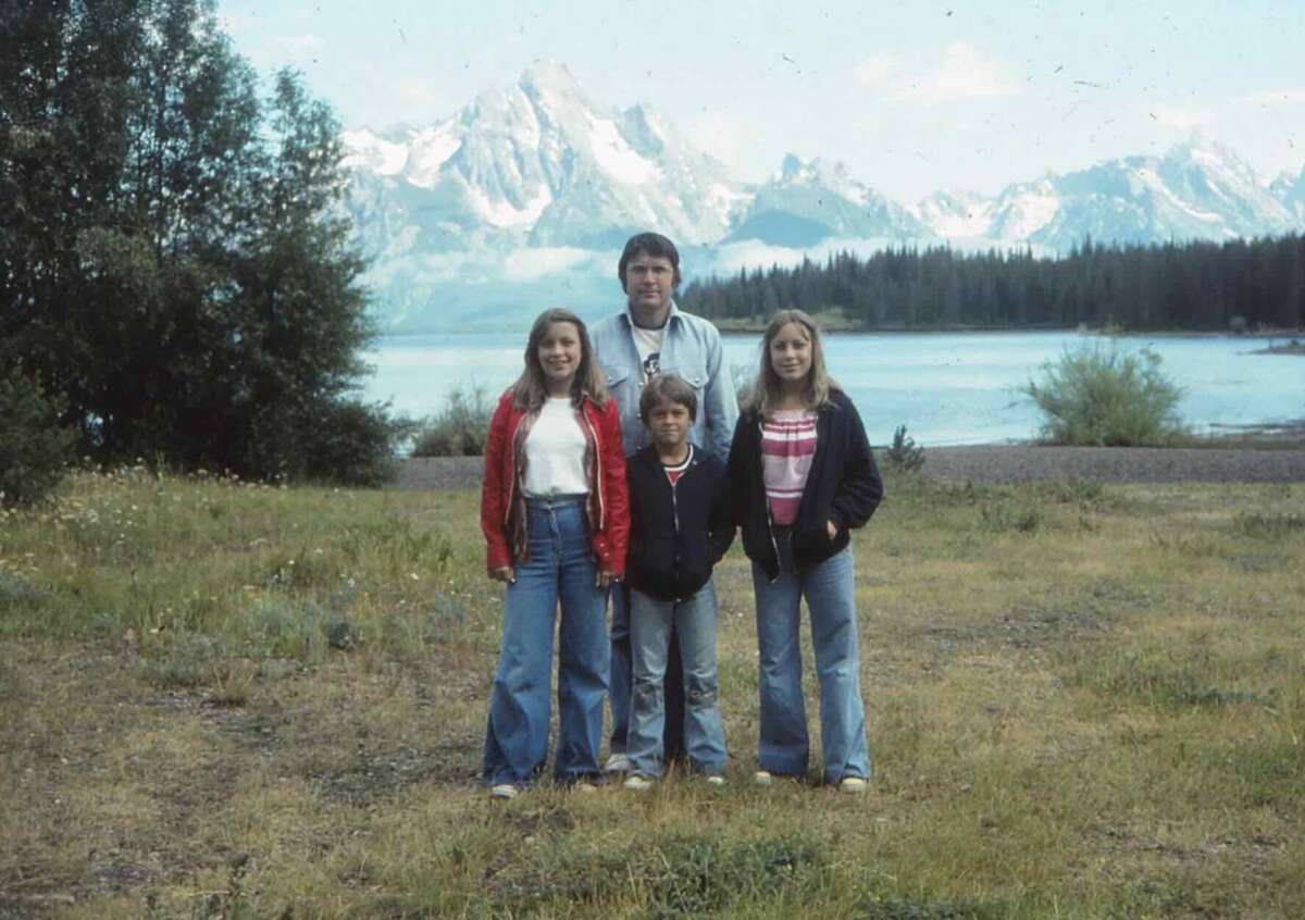 A man and three children stand together on grass near a lake, with snow-capped mountains and trees in the background. The group is wearing casual clothes and jackets, posing for the photo outdoors.