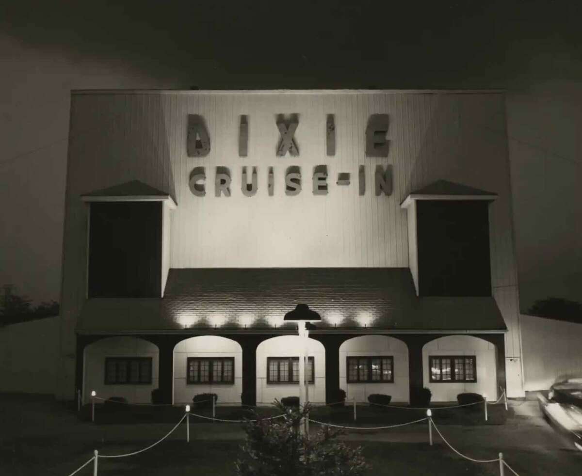 A black-and-white photo of the Dixie Cruise-In drive-in theater at night, with bold illuminated letters on the building and small lights lining the walkway to the entrance.