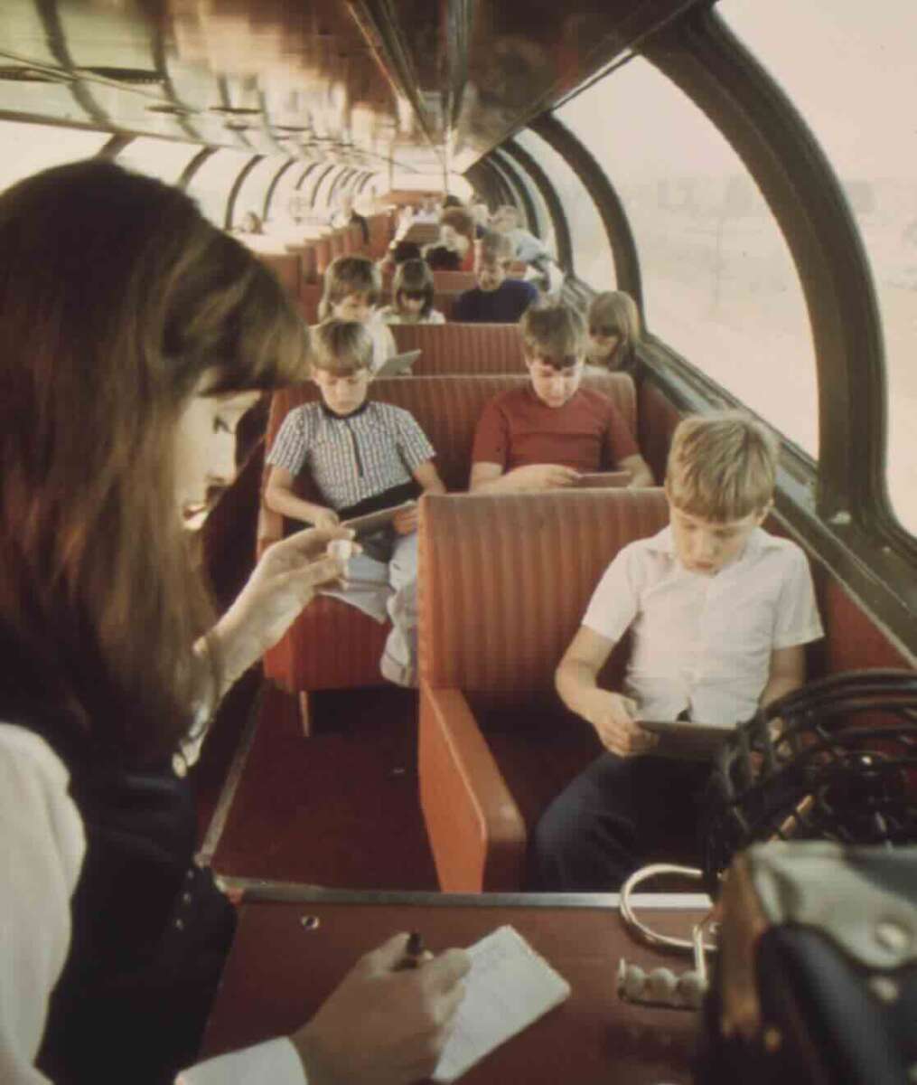 Children sit and read or write in a spacious, retro train car with large windows. A woman in the foreground takes notes, while sunlight streams through the curved glass roof. The atmosphere feels studious and relaxed.