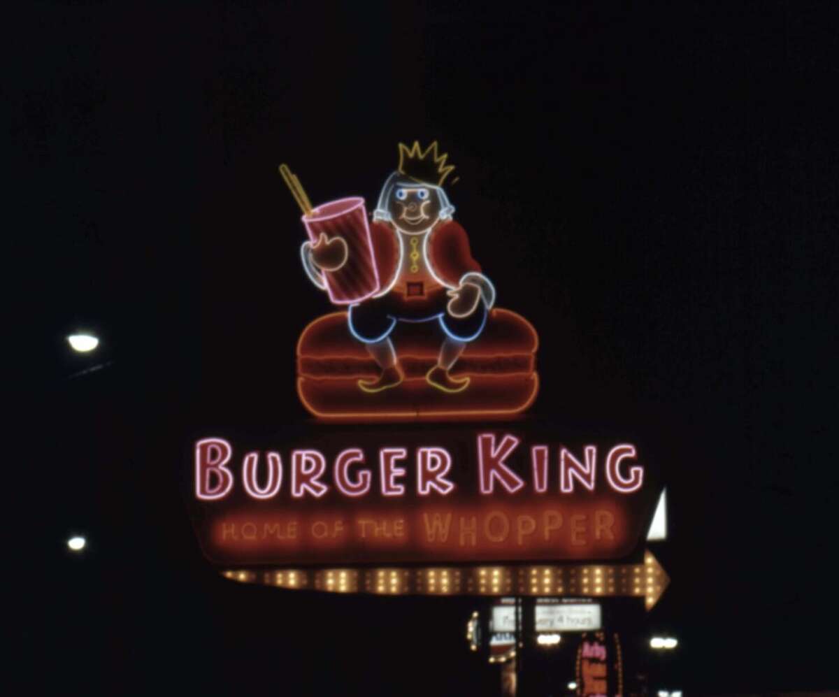 A neon Burger King sign at night shows a cartoon king with a crown holding a drink, sitting on a burger. The text reads "Burger King Home of the Whopper.