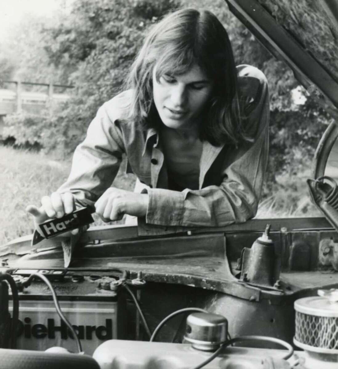 A person with long hair and an open shirt is working under the hood of a car, holding a DieHard battery, outdoors with trees in the background.