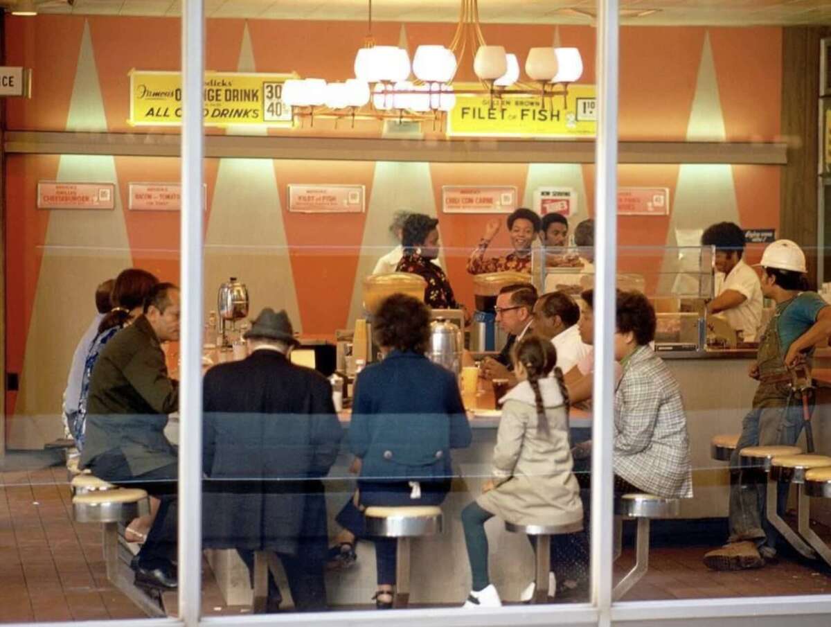 A group of people sit at a retro-style diner counter with orange decor, enjoying food and drinks. Vintage signs and hanging lights are visible, creating a nostalgic atmosphere. The scene is viewed from outside through large windows.