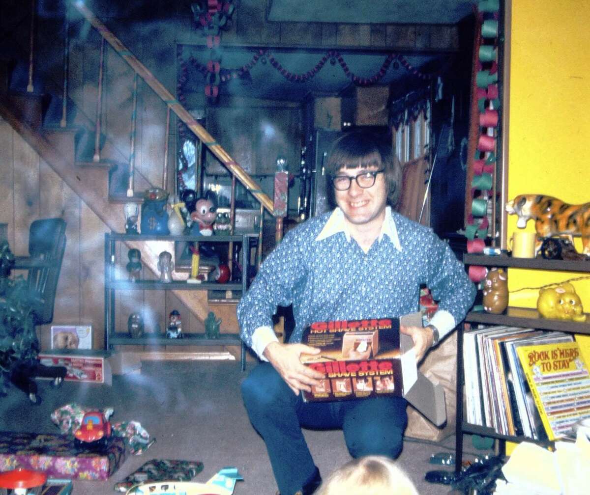 A man with glasses and a patterned blue shirt smiles while holding a boxed gift in a warmly lit living room with wood paneling, colorful paper chains, toys, and books on shelves in the background.