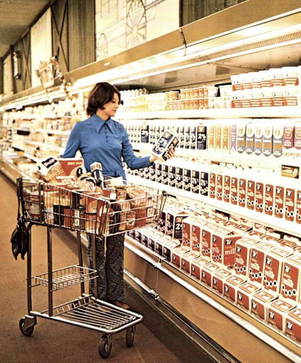 A woman in a blue shirt shops for milk and dairy products in a supermarket aisle, pushing a metal cart filled with groceries. Shelves are stocked with various milk cartons and other dairy items.