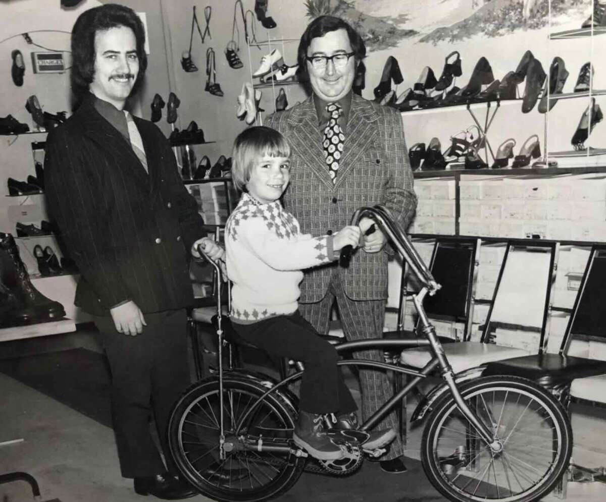 A black-and-white photo shows a child smiling while sitting on a bicycle, flanked by two men in suits. They are indoors, surrounded by rows of shoes displayed on shelves and walls.