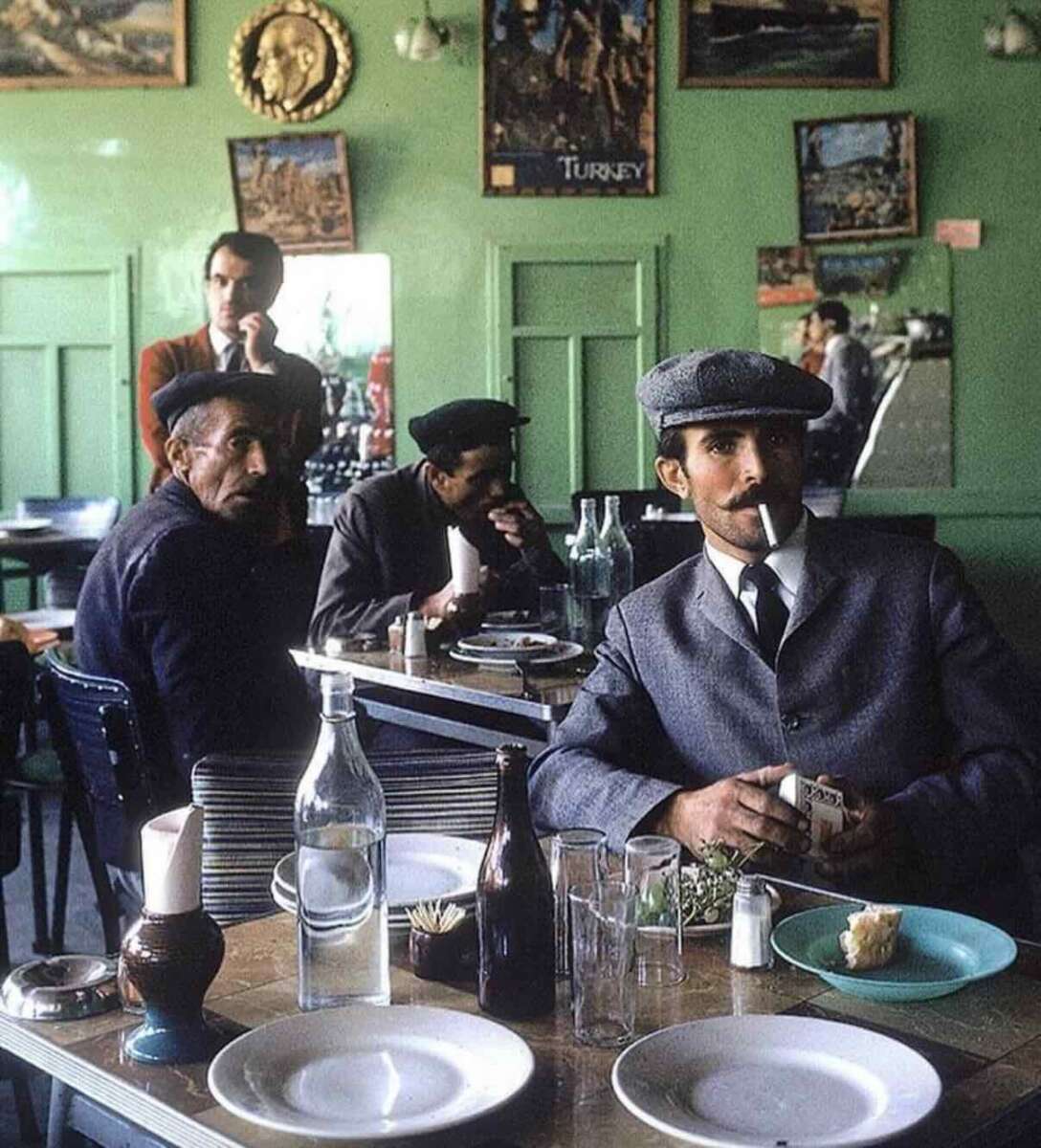 Four men in vintage clothing sit in a green-walled cafe. One man in front, wearing a cap and suit, smokes a pipe and holds a cigarette pack. Food, bottles, and glasses are on the table. Framed pictures decorate the wall.