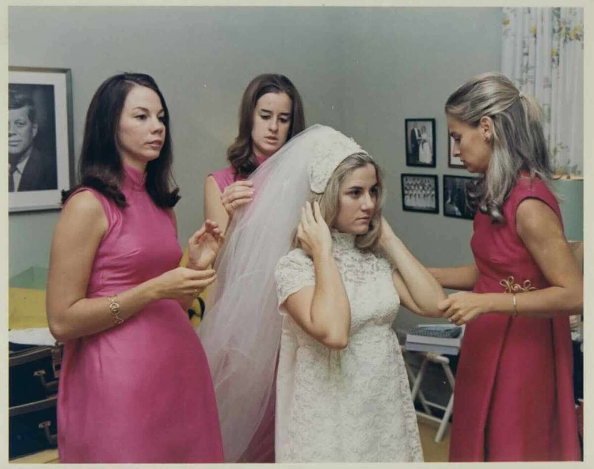 A bride in a white lace dress and veil is being helped by three women in matching pink dresses. They are in a room with framed photos on the wall and a portrait of John F. Kennedy in the background.
