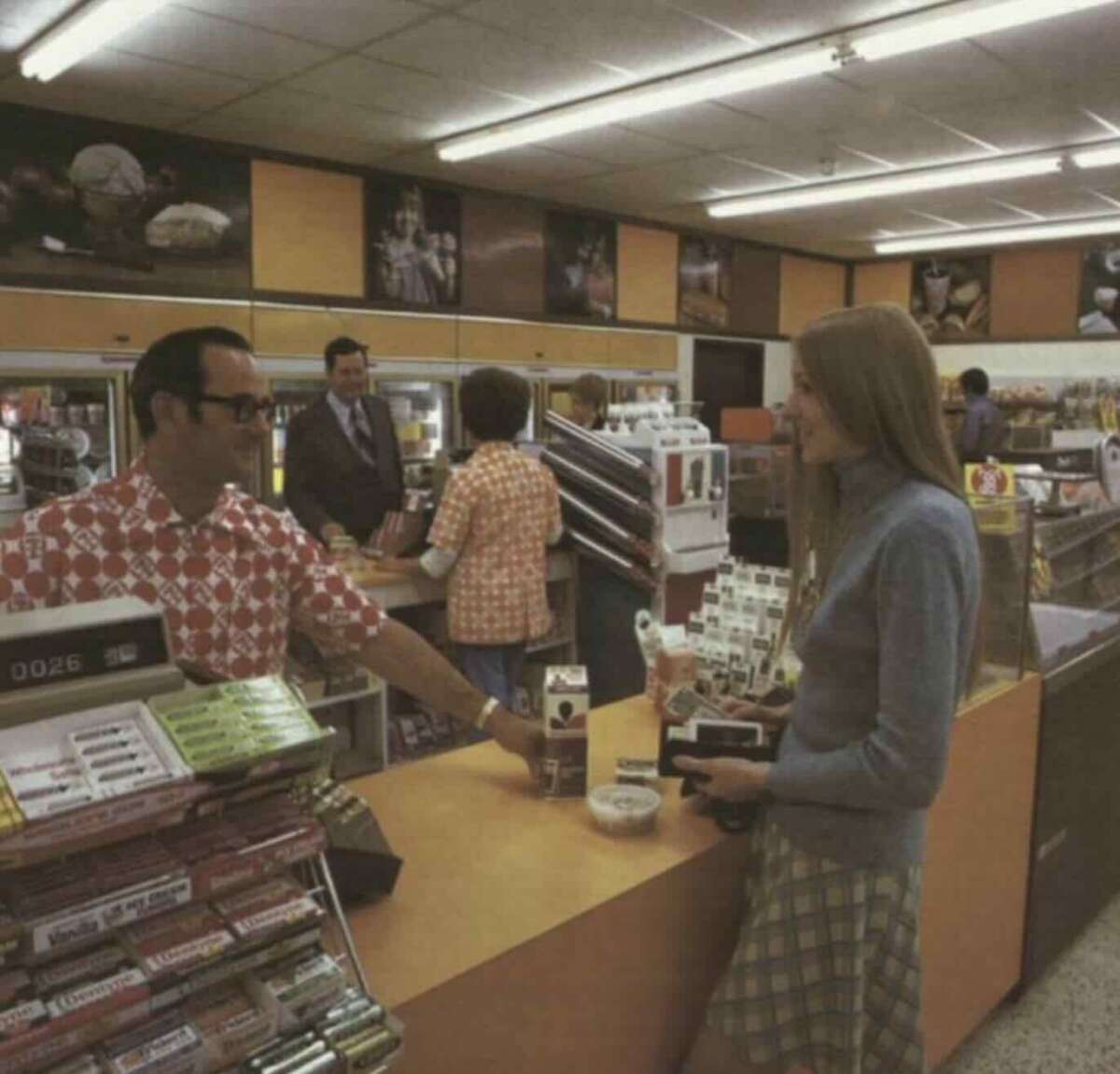 A woman with long hair pays at a convenience store counter, holding a wallet and camera. A male cashier in glasses and a patterned shirt assists her; other customers and shelves of snacks are visible in the background.