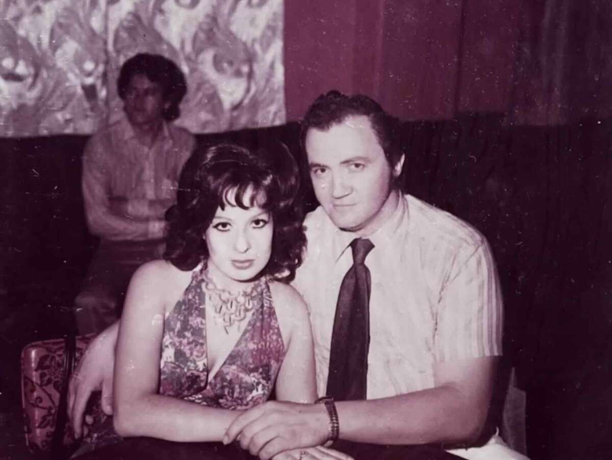 A vintage photo shows a woman in a floral dress and a man in a striped shirt and tie sitting close together, posing for the camera. Another man sits in the background against a patterned wall.