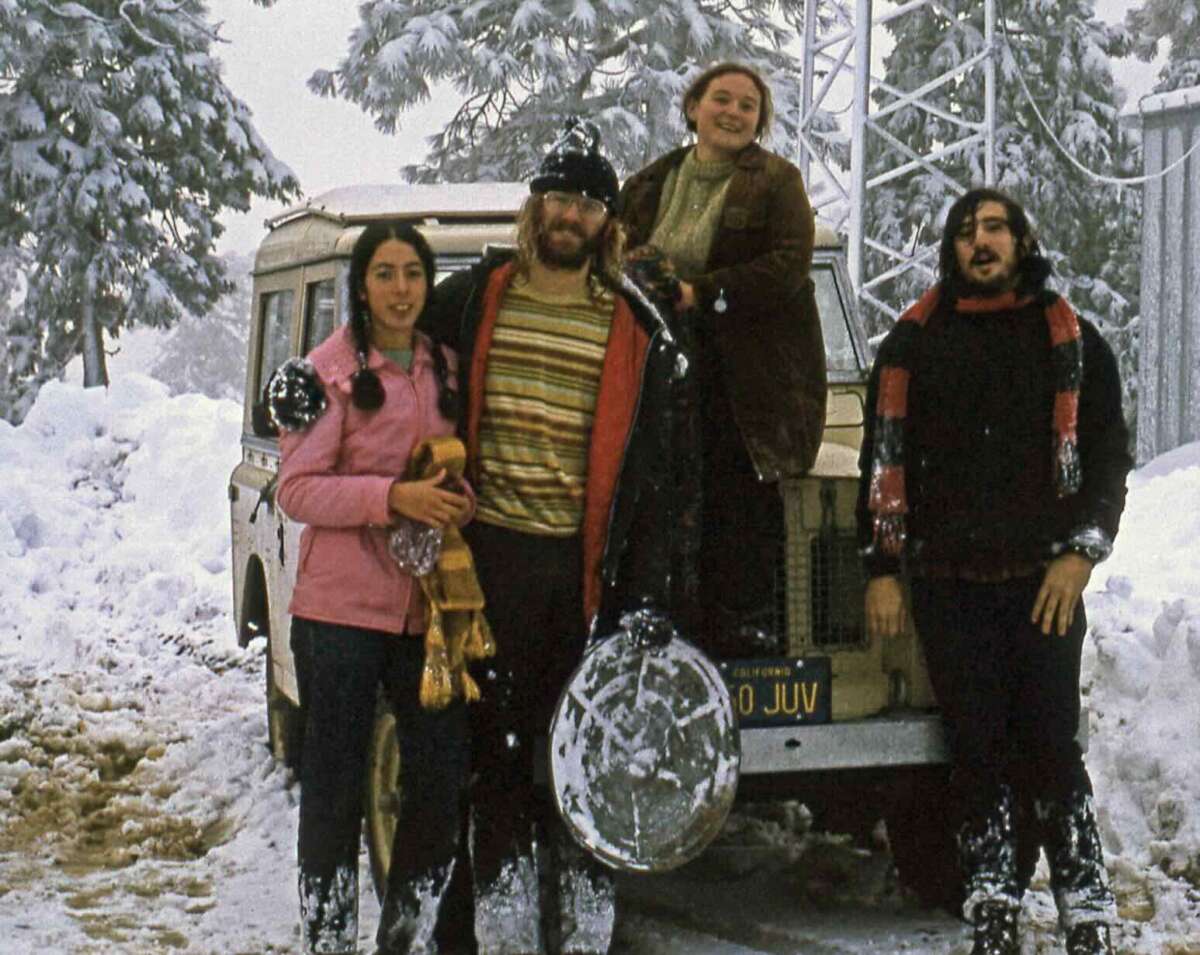 Four people in winter clothes stand in front of a snow-covered vehicle, surrounded by deep snow and trees. Some of them have snow on their clothes and appear to be smiling and enjoying the snowy environment.
