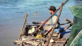 A man in a hat sits on a homemade wooden raft by a river, flipping food in a frying pan over a camp stove, with cooking supplies and logs visible around him.