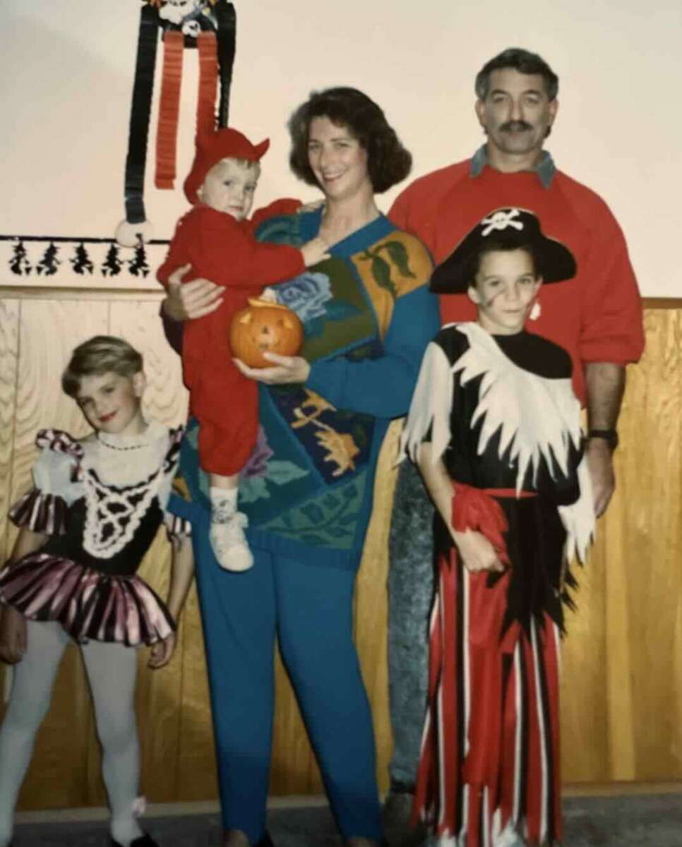 A family poses for a photo in Halloween costumes. A woman holds a toddler in a devil costume with a pumpkin; a man stands behind them. Two children are dressed as a ballerina and a pirate. Halloween decorations hang on the wall.