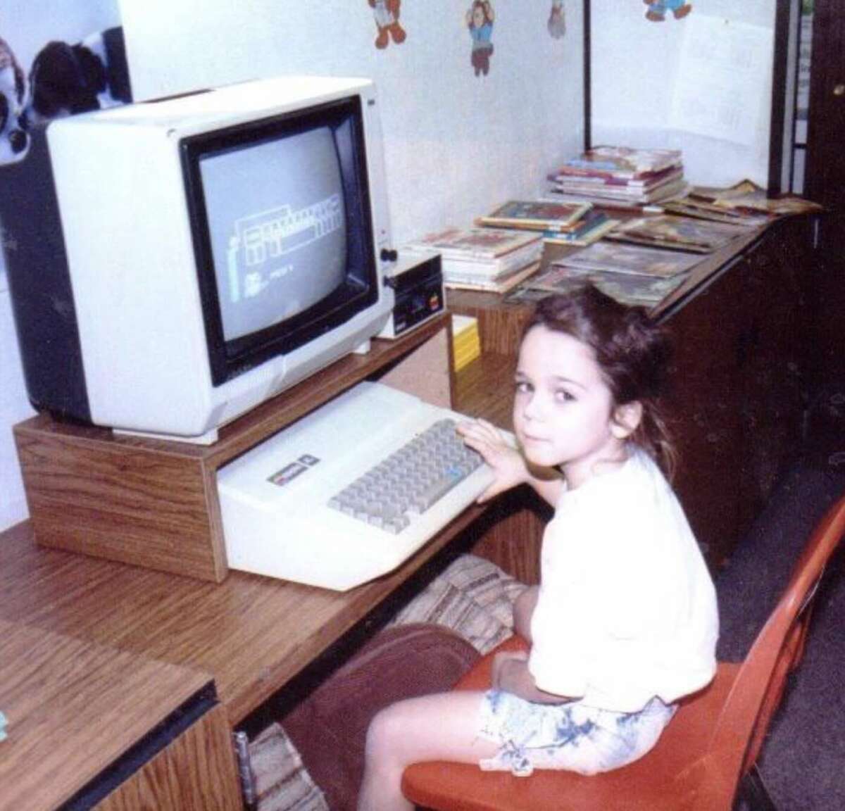 A young girl sits at a desk using a vintage computer with a CRT monitor. She looks toward the camera. Books and papers are stacked on the desk, and cartoon decorations are on the wall behind her.
