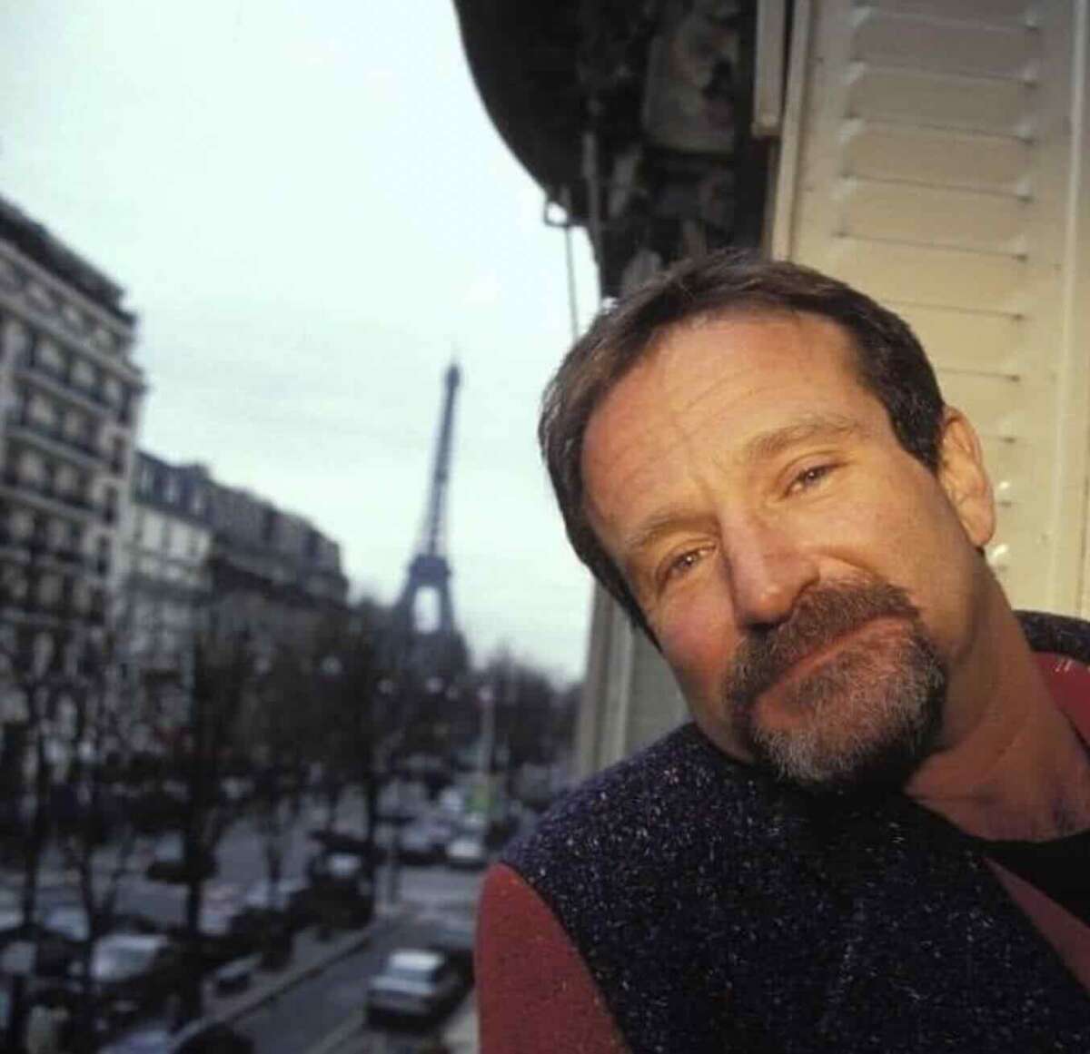 A man with a short beard leans against a window ledge, smiling softly. In the background, a street scene and the Eiffel Tower are visible, suggesting the photo was taken in Paris.