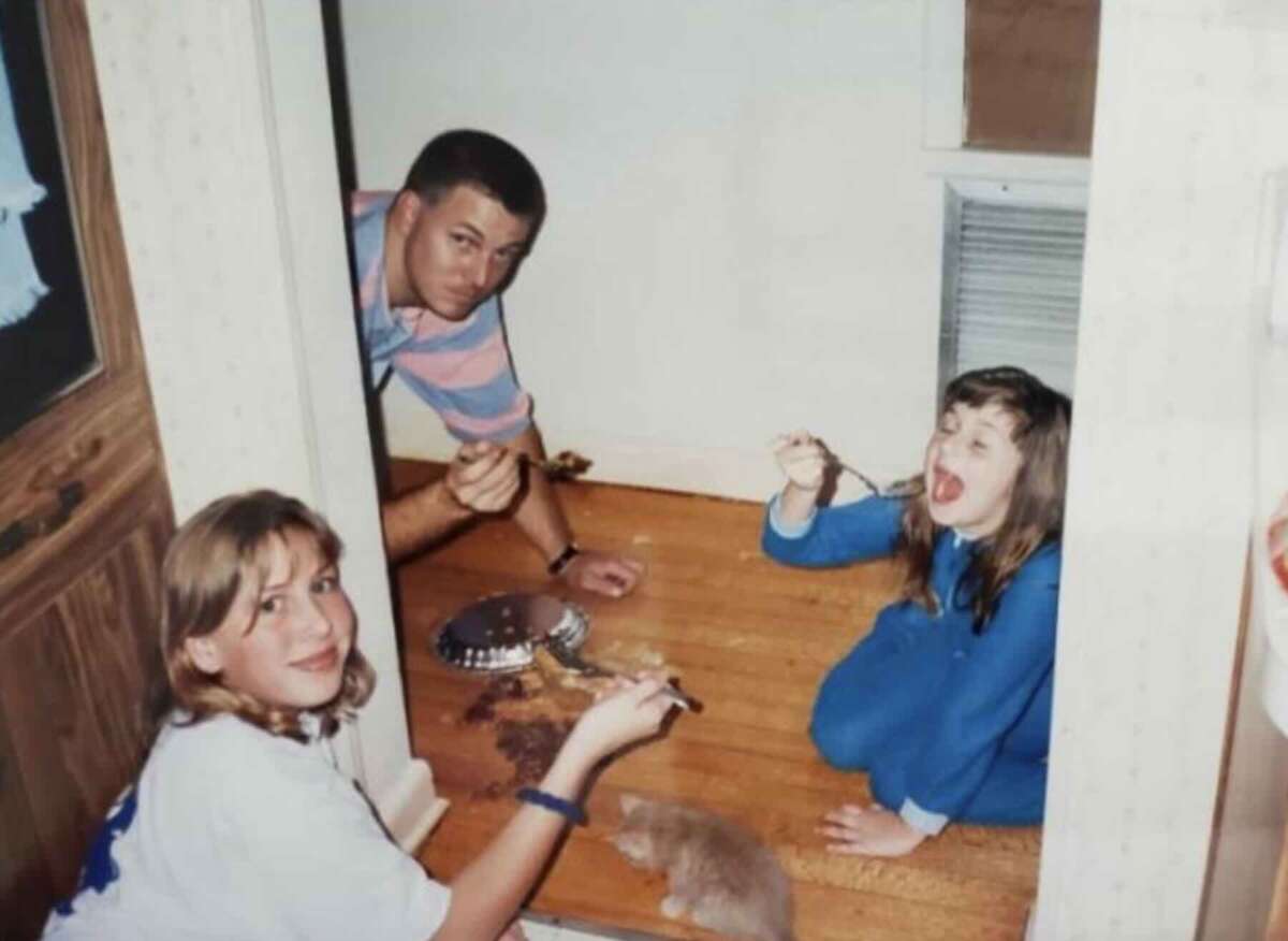A man and two girls sit on a wooden floor, eating chocolate cake directly from a pan with spoons. The younger girl, in blue pajamas, looks excited. A small cat or kitten is near the pan.