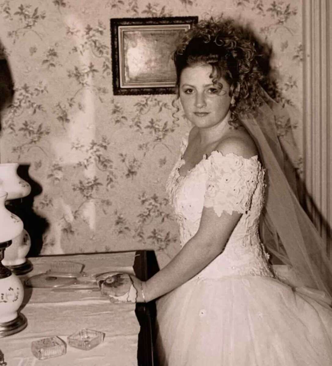 A bride in a white wedding dress with lace details and a veil poses indoors by a table with a vintage lamp and ashtrays, against a floral wallpapered wall.