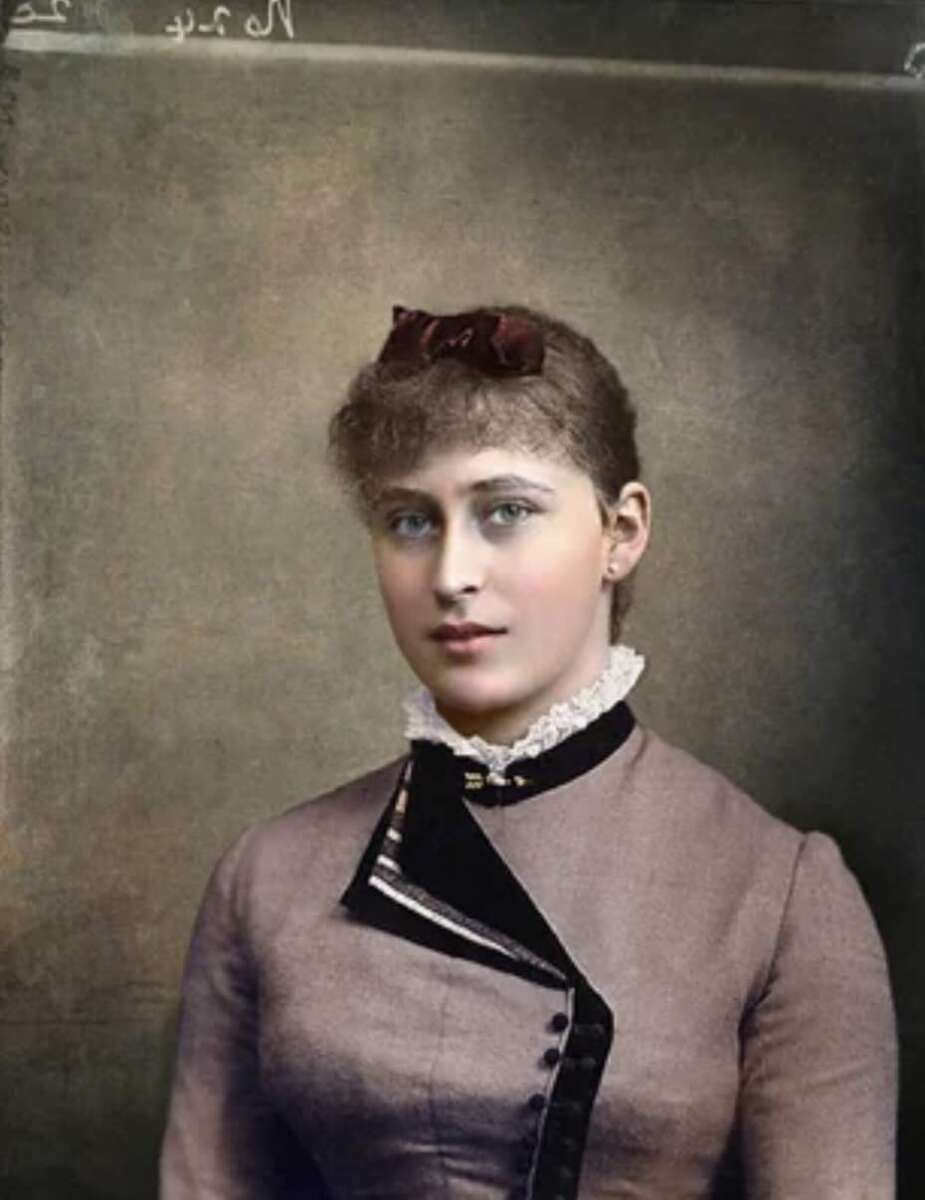 A young woman with light skin and blue eyes wears a high-collared, Victorian-style dress with lace trim and a dark ribbon at the neck. Her brown hair is styled up with a bow, and she sits in front of a plain studio backdrop.