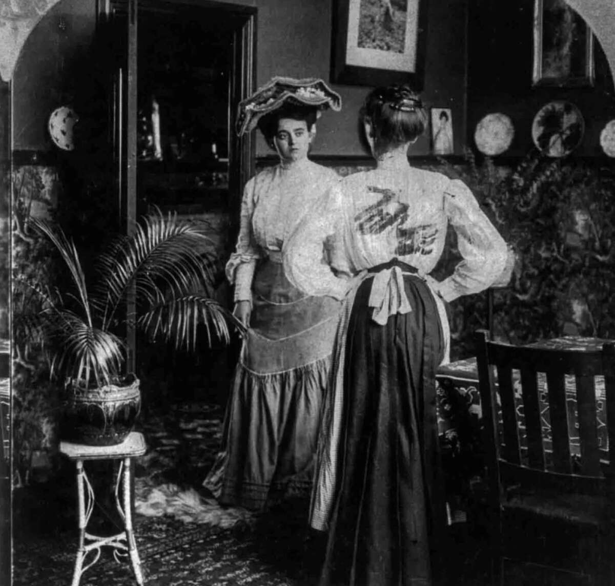 Two women in early 1900s attire stand in a richly decorated room. One faces the camera, wearing a large hat, while the other has her back turned. The room features patterned wallpaper, framed art, and potted plants.
