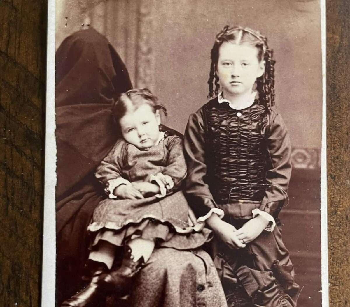 A vintage photograph of two young girls in Victorian-style dresses. The older girl stands with hands clasped, while the younger sits on a lap, likely of a hidden adult draped in dark fabric, creating a ghostly effect.