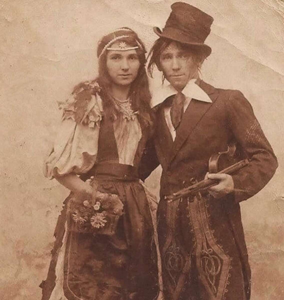 A sepia-toned vintage photograph of two people in old-fashioned attire. The person on the left wears a floral dress and headband, holding flowers. The person on the right wears a suit, top hat, and cravat, holding a violin.