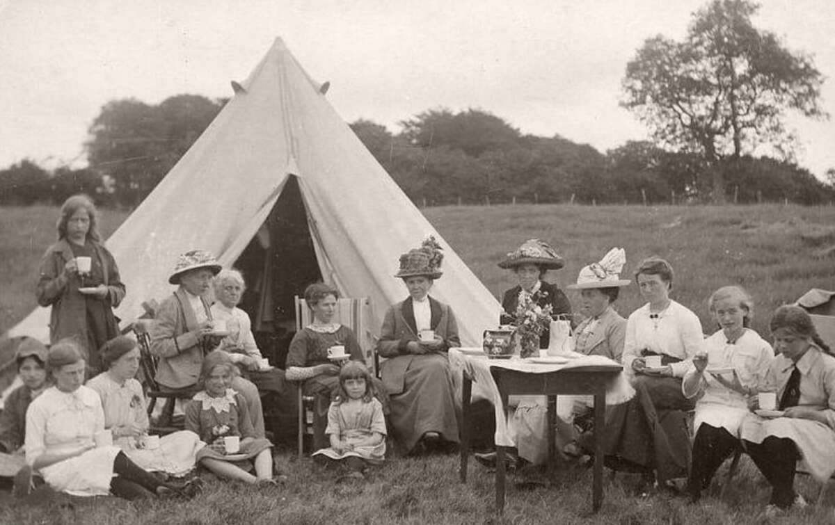 A group of women and children in vintage clothing sit and stand around a table with tea outdoors in front of a large canvas tent, enjoying a tea party in a grassy field.