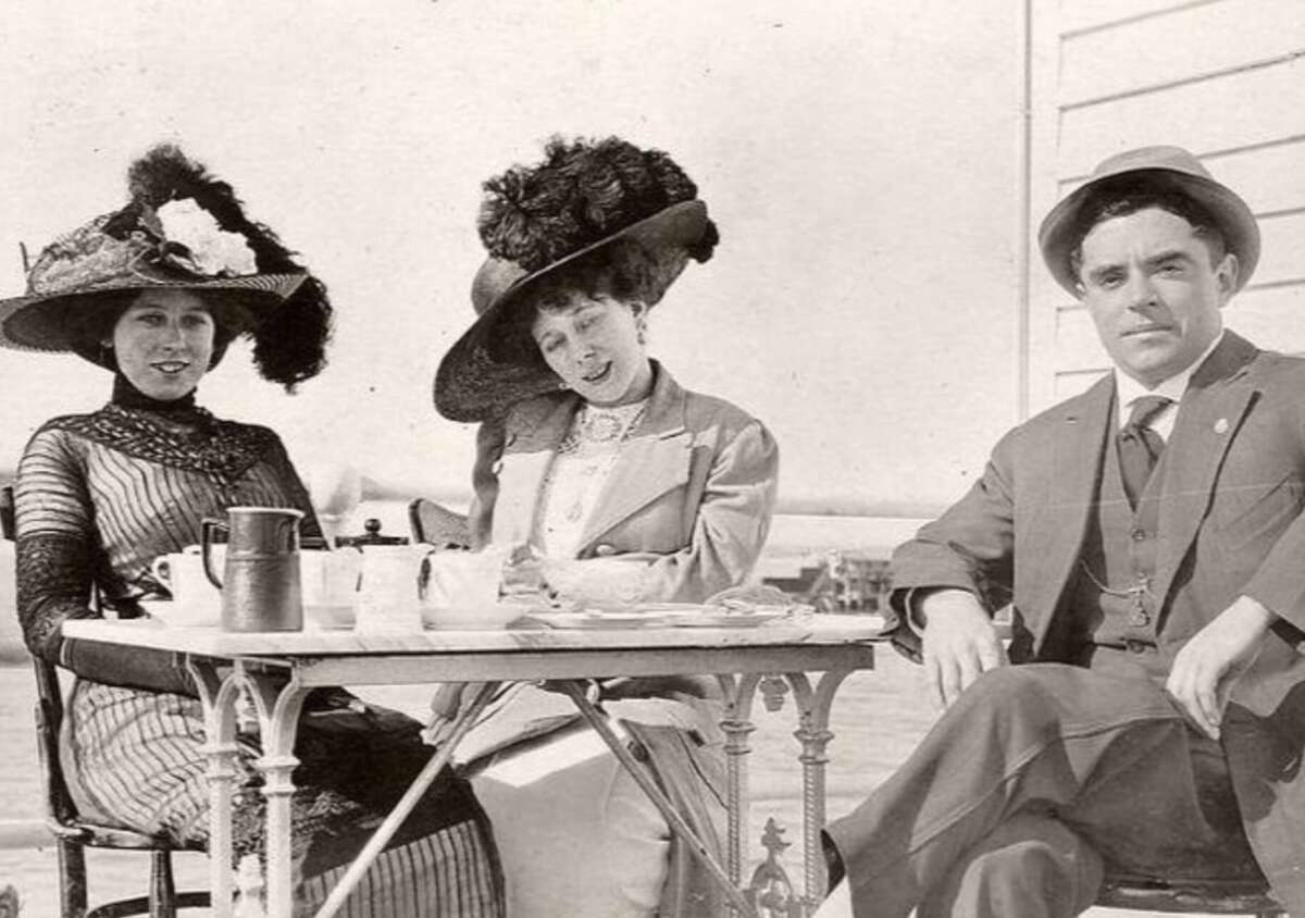Three people in early 1900s attire sit at an outdoor table with tea cups and a teapot. Two women wear large decorative hats and dresses, and a man in a suit and hat sits beside them.