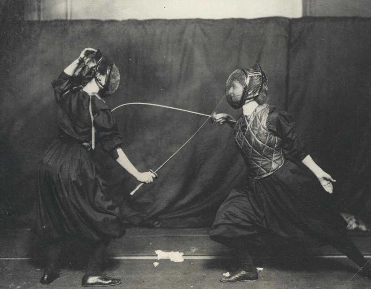 Two women in protective gear and masks engage in a fencing duel indoors, both holding swords with one lunging forward and the other preparing to block. The scene is in black and white.
