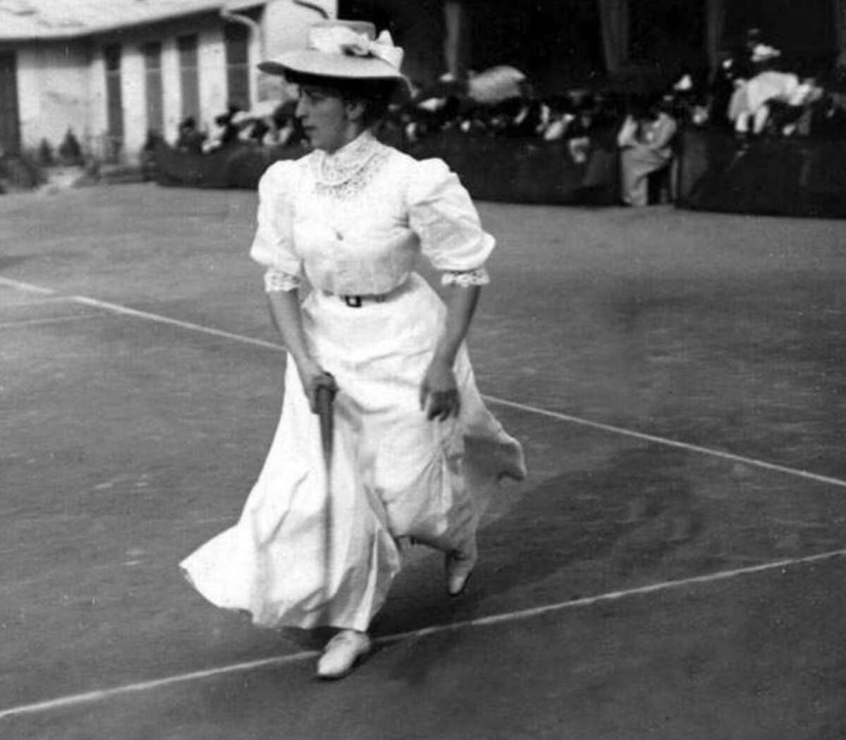 A woman in a long, white Victorian-era dress and hat holds a tennis racket on an outdoor court, with spectators seated in the background.