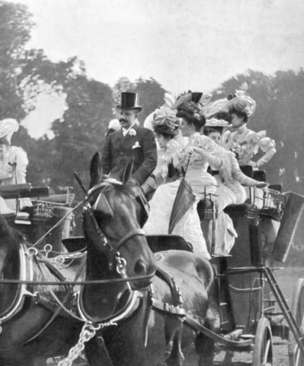A black-and-white photo of elegantly dressed women and a man in Victorian-era attire riding in a horse-drawn carriage, surrounded by trees. The women wear ornate hats and dresses, and the man wears a top hat and suit.