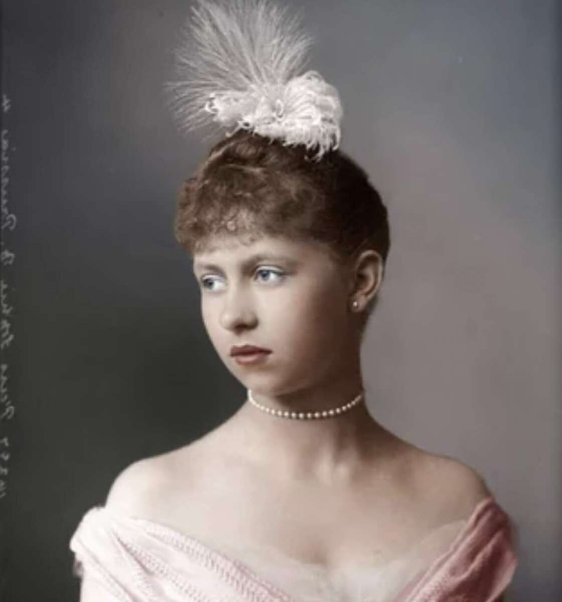 A young woman in a pink off-shoulder gown, wearing a pearl necklace and a feathered hair accessory, looks to the side with a calm expression against a muted background.