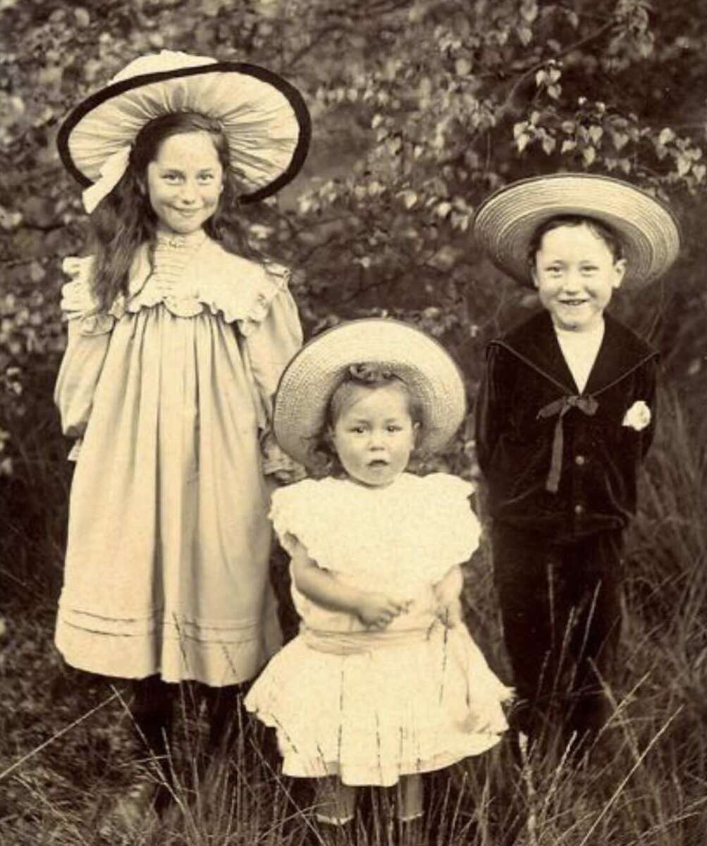Three children in vintage clothing and large hats pose outdoors. The older girl and boy stand on either side of a younger girl in a white dress. Bushes and grass fill the background, giving a natural, old-fashioned scene.