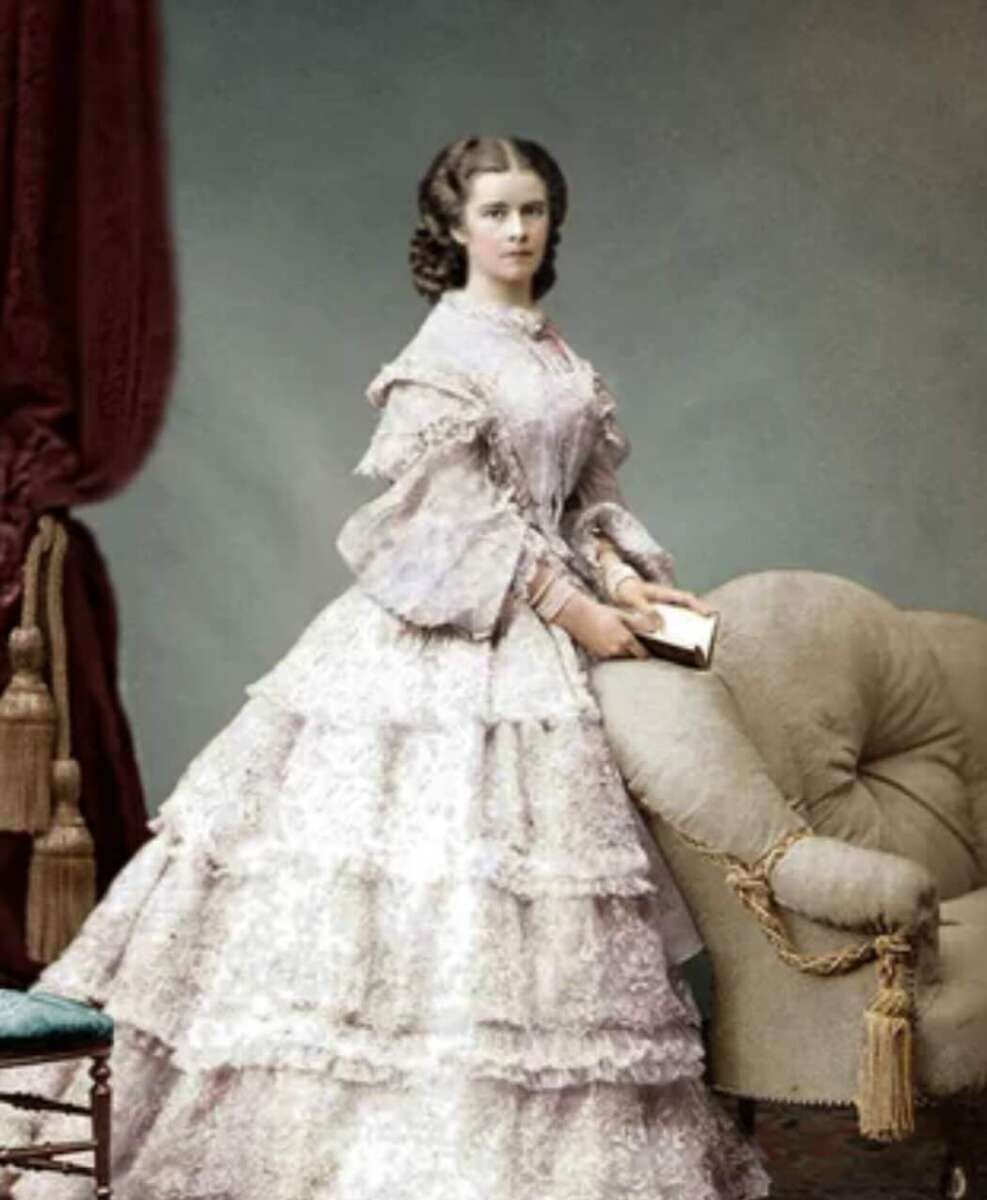 A woman in a formal, ruffled, light-colored Victorian-era dress stands beside an ornate armchair, holding a closed fan. The background features a red curtain and a small part of a blue-cushioned chair.