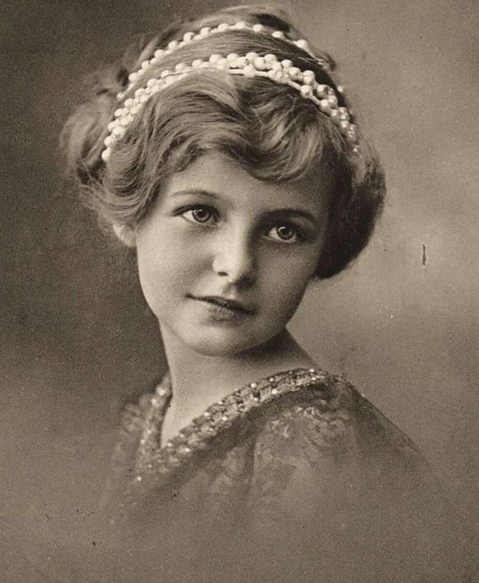 A young woman with curled light hair, adorned with a beaded headband, gazes softly at the camera. She wears a decorative, vintage-style dress. The photo is in sepia tones, giving it an antique feel.
