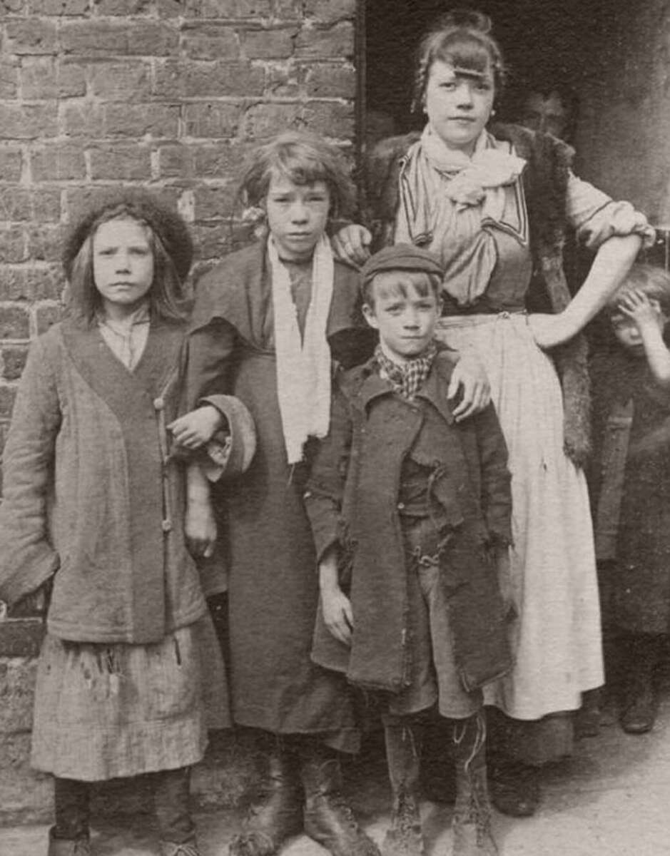 Four children in worn, old-fashioned clothing stand outside a brick building. Three are in front; a girl and a boy stand beside an older girl, who has her arm around them. Another child peers from behind her.