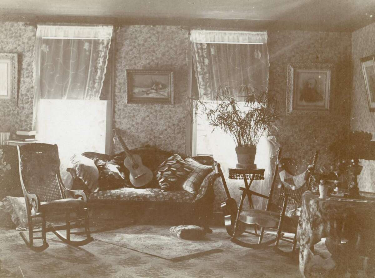 A sepia-toned photo of a vintage living room with patterned wallpaper, two windows with sheer curtains, a couch with pillows and a guitar, rocking chairs, paintings on the wall, and potted plants on tables.