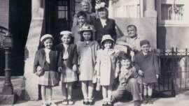 A black-and-white photo of a group of children and adults, dressed in vintage clothing, posing and smiling on the steps of a building with columns and a railing, likely taken in the mid-20th century.