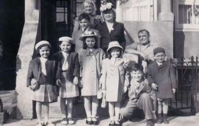 A black-and-white photo of a group of children and adults, dressed in vintage clothing, posing and smiling on the steps of a building with columns and a railing, likely taken in the mid-20th century.