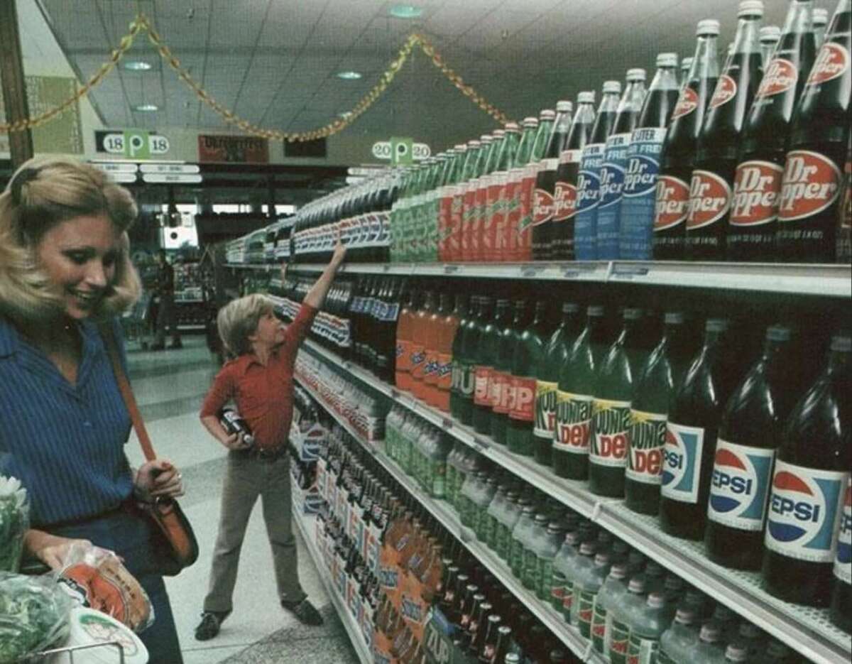A woman pushes a shopping cart while a boy reaches for a bottle on a grocery store shelf lined with various soda brands, including Dr Pepper, 7UP, Mountain Dew, and Pepsi.