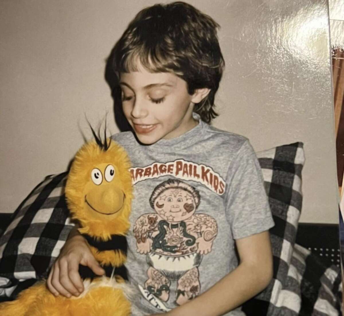 A smiling child with short brown hair sits on a plaid-patterned cushion, wearing a Garbage Pail Kids T-shirt and holding a large, plush yellow bee toy.