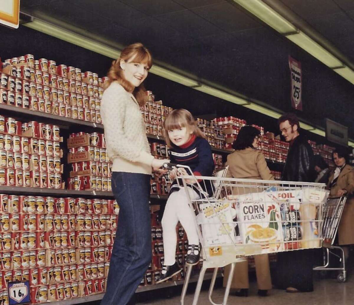 A woman smiles while holding a child sitting in a grocery cart in a supermarket aisle lined with stacked cans and a box of corn flakes in the cart. Other shoppers browse in the background.