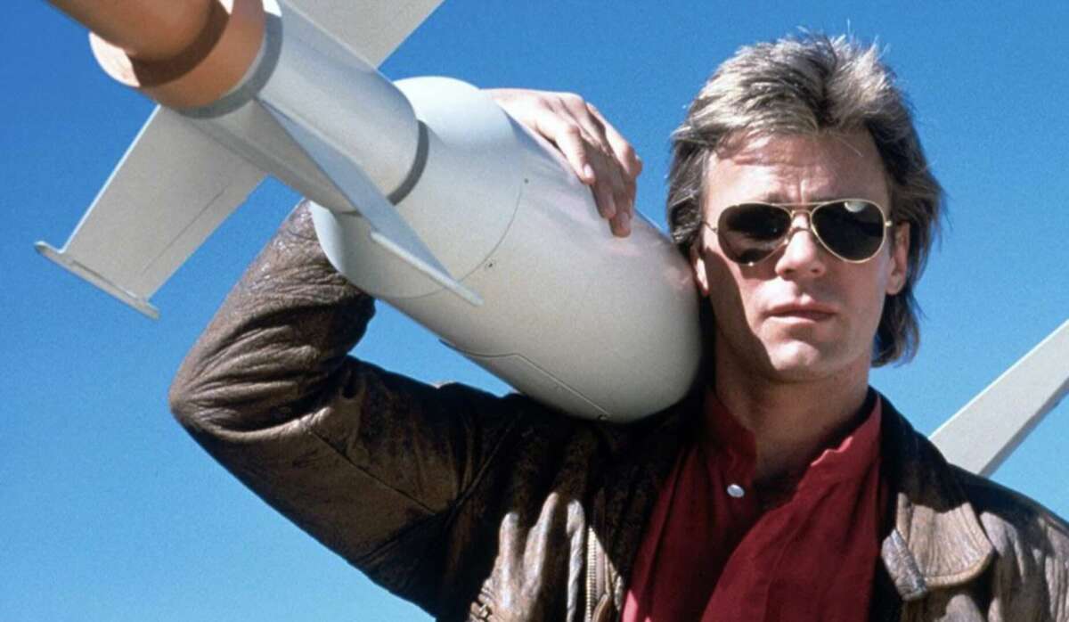 A man wearing sunglasses and a brown leather jacket holds a large missile over his shoulder, standing against a clear blue sky.