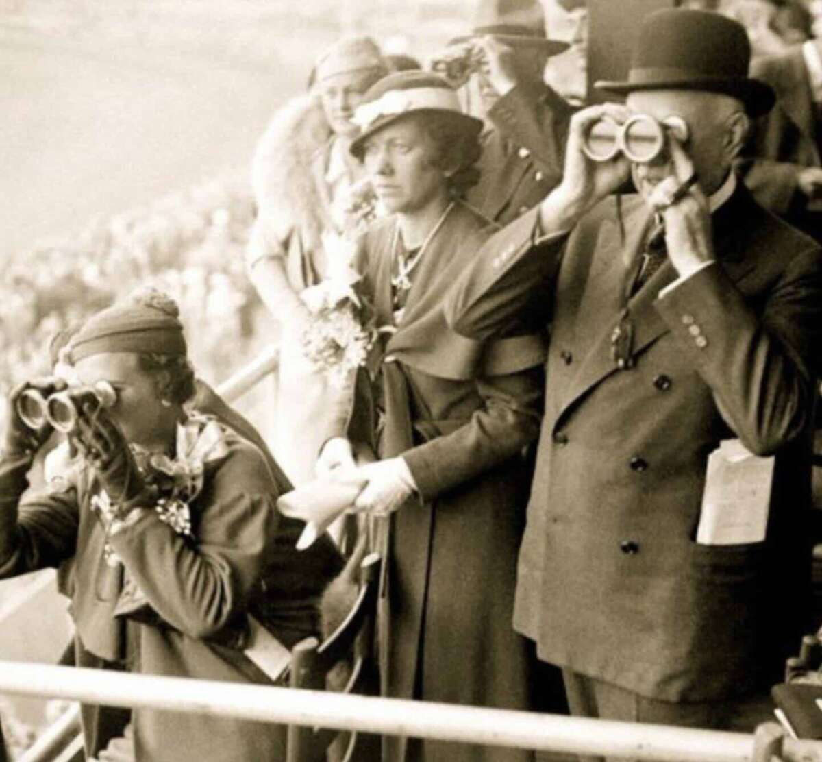 A vintage black-and-white photo shows three elegantly dressed people at an outdoor event; two use binoculars, while the woman in the middle stands without them, holding her gloves and looking ahead.