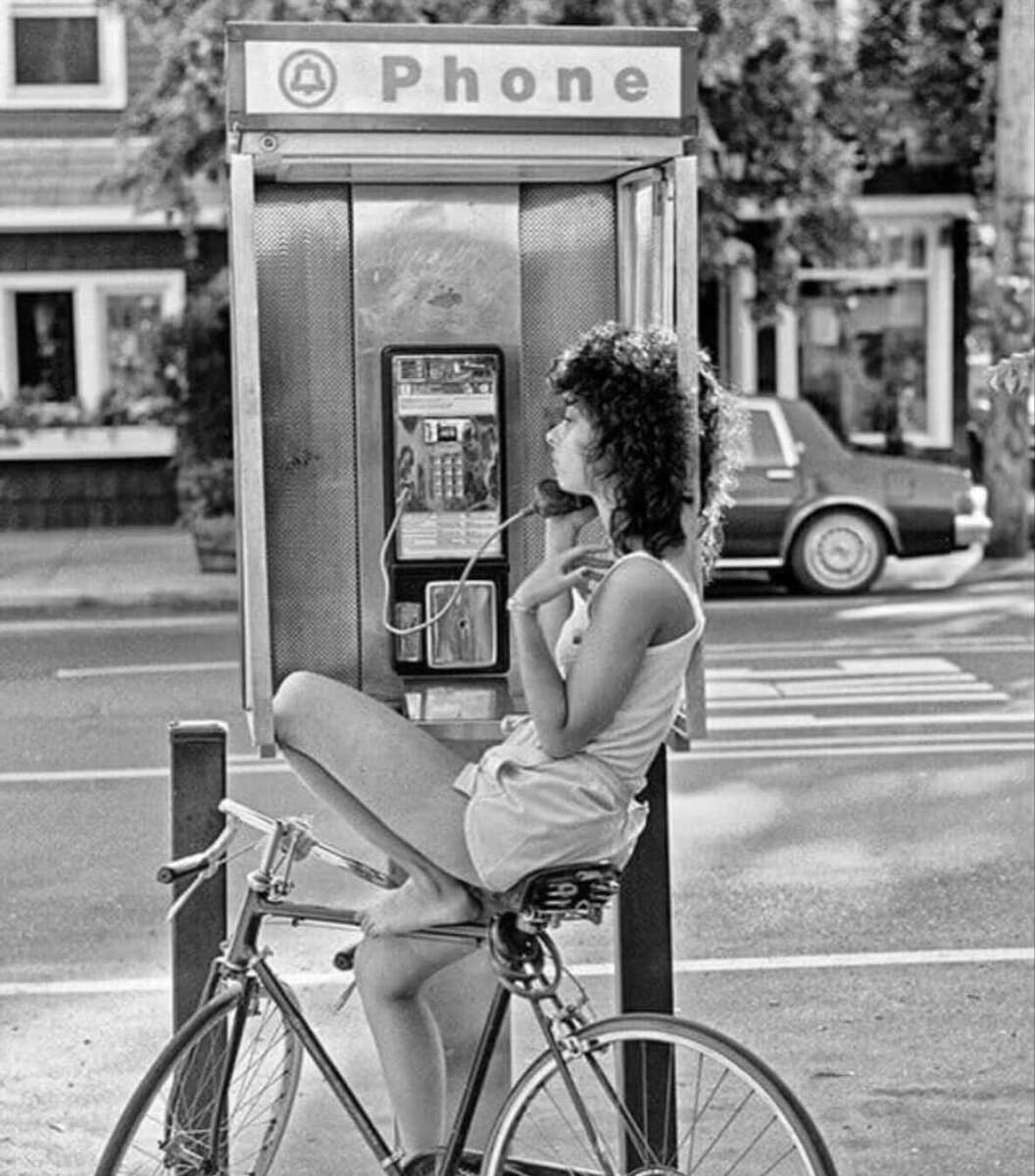 A woman with curly hair in shorts and a tank top sits on a bicycle, using a payphone on a city sidewalk. A car and houses are visible in the background. The image is black and white.